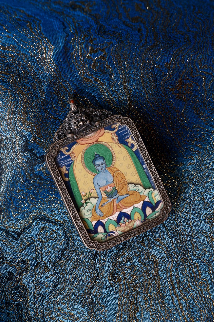 Flat lay of Medicine Buddha thangka pendant in vintage silver gawu case on blue brocade textile background