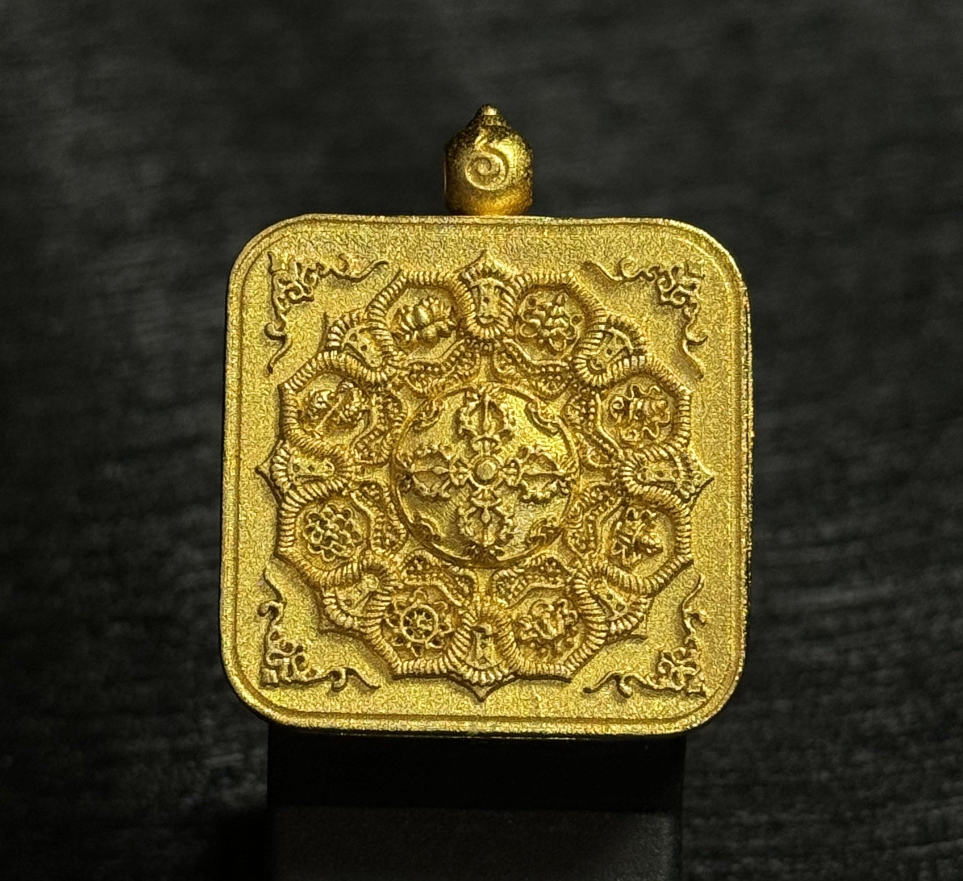 Back side of a Tibetan miniature thangka pendant featuring a raised mandala relief design engraved on a textured gold-toned metal surface, with traditional decorative motifs symbolizing sacred protection and Buddhist symbolism.