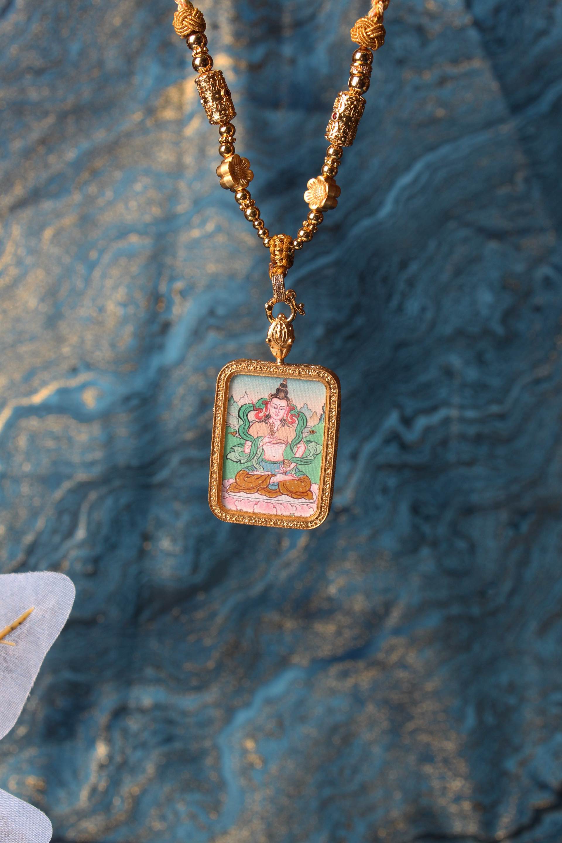 Tibetan Vajrasattva mini thangka pendant shown with gold-tone beads and frame, hand-painted mineral pigment artwork, decorative background setting