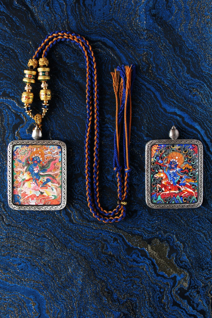 Flat-lay showing two Palden Lhamo mini thangka pendants side by side, each framed in a silver protective case, with a blue-and-gold braided cord necklace and loose tassel cords on a deep blue brocade background