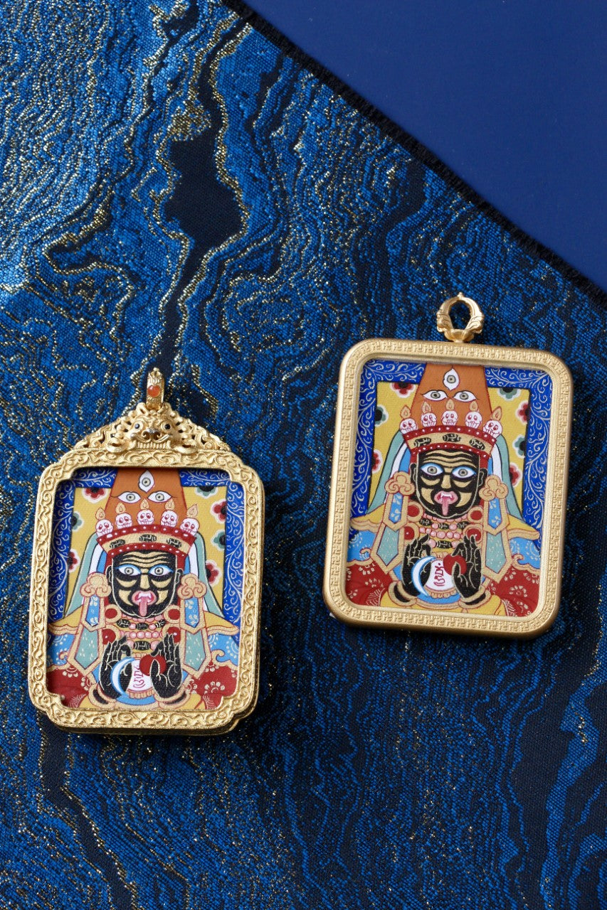Two similar Zaki Lhamo mini thangka pendants side by side, comparing an ornate gawu-style frame and a classic square gold-tone copper frame on blue textile