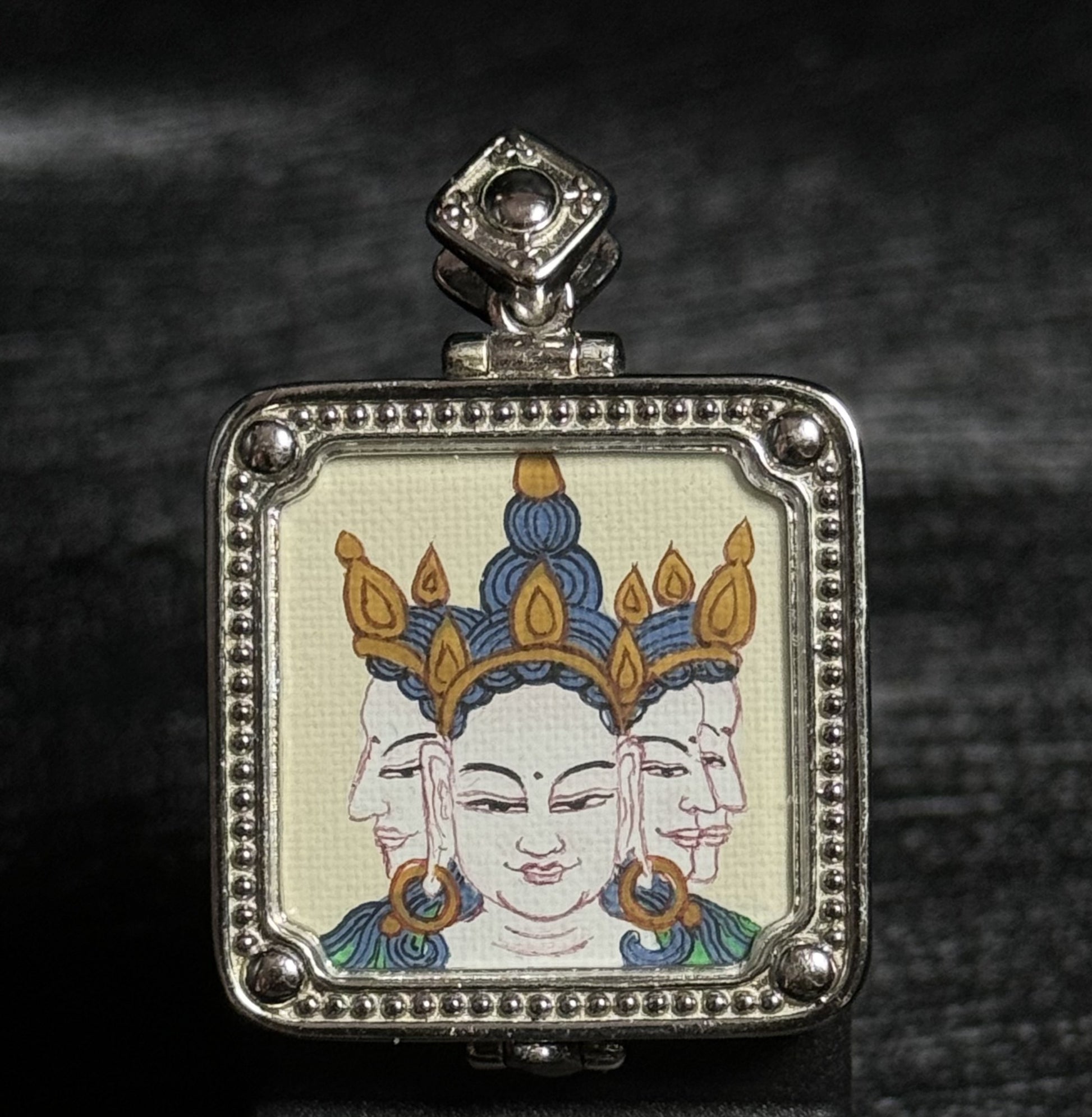 Front view of a hand-painted Vairocana Buddha miniature thangka pendant showing the three-faced Buddha depiction painted with mineral pigments inside a detailed 925 silver plated frame.