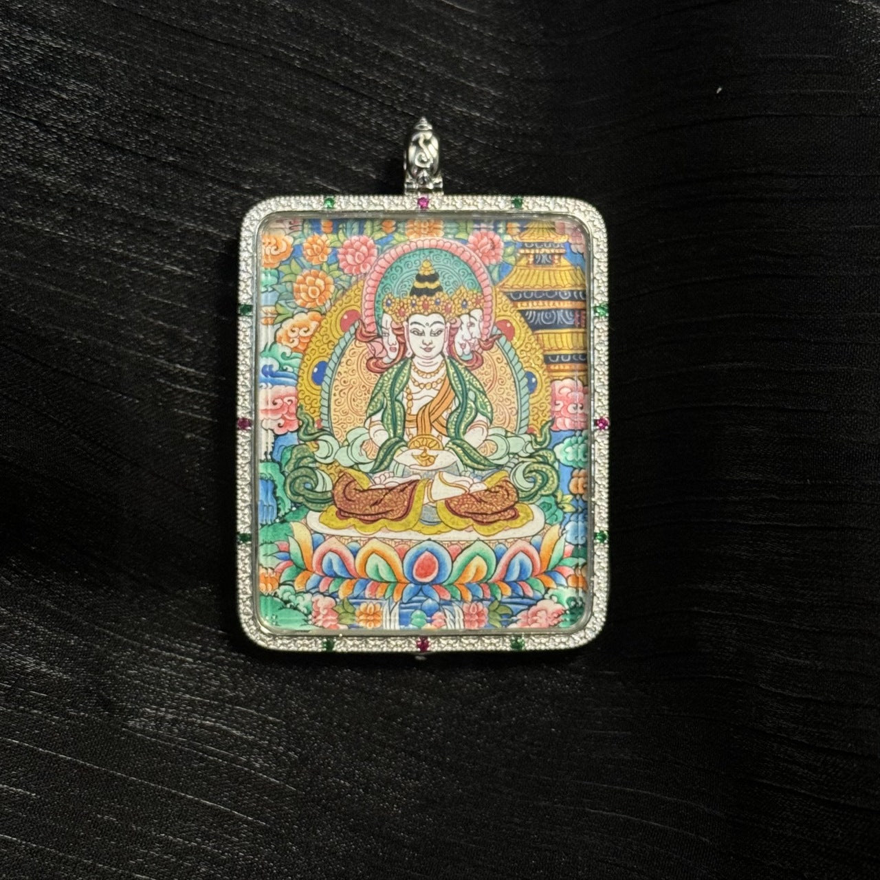 Close-up front view of the Vairocana Buddha mini thangka pendant featuring the serene Buddha seated on a lotus throne surrounded by colorful floral motifs and traditional Tibetan decorative elements within a silver pendant frame.