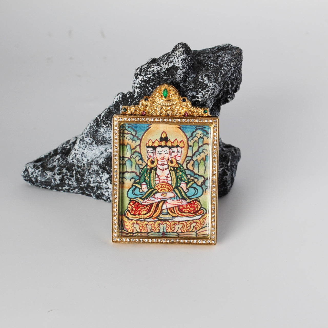 Front view of Vairocana Buddha mini thangka pendant showing the hand-painted multi-faced Vairocana figure with halo, gold-tone rectangular frame with crystal border, and ornate crown top with a green gemstone, photographed on a rock prop.