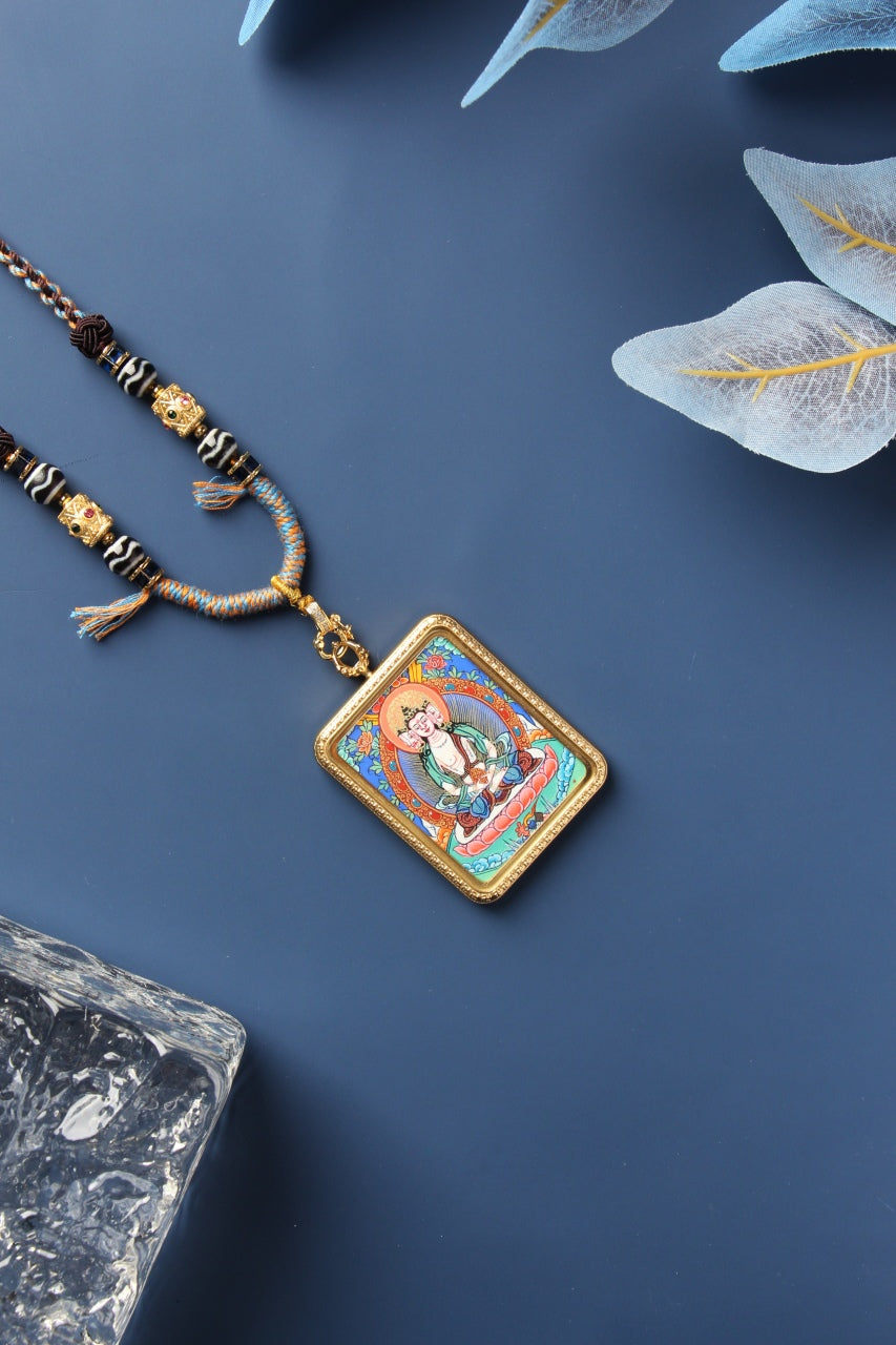 Vairocana Buddha mini thangka pendant necklace in a gold-tone titanium steel case, photographed diagonally on a matte blue background with braided cord and decorative beads.