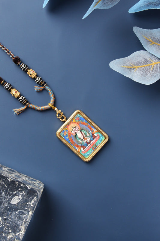 Vairocana Buddha mini thangka pendant necklace in a gold-tone titanium steel case, photographed diagonally on a matte blue background with braided cord and decorative beads.