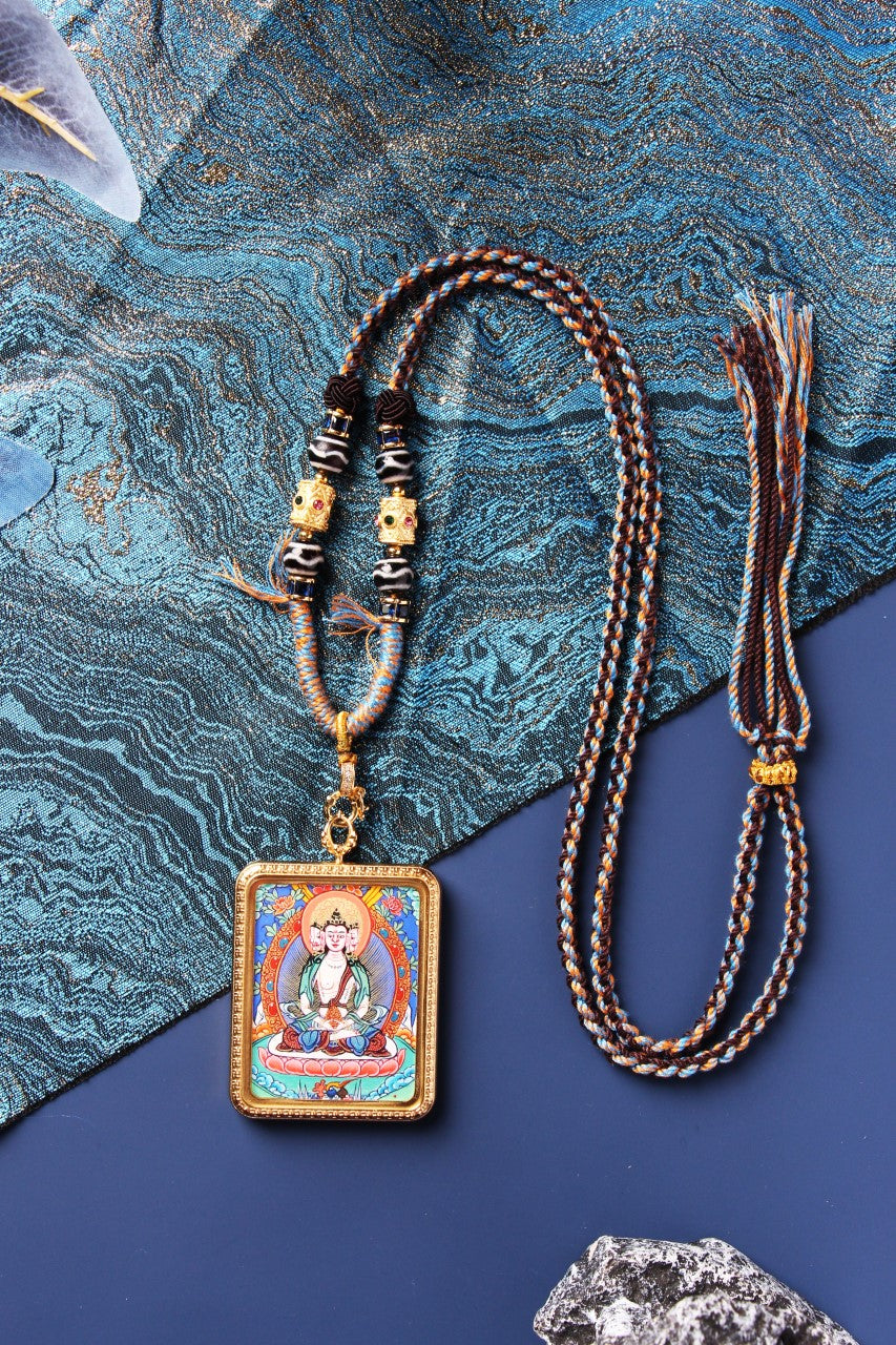 Full view of a Vairocana Buddha mini thangka pendant necklace with braided cord, bead details, and an additional matching cord, styled on a blue textured cloth and dark background.