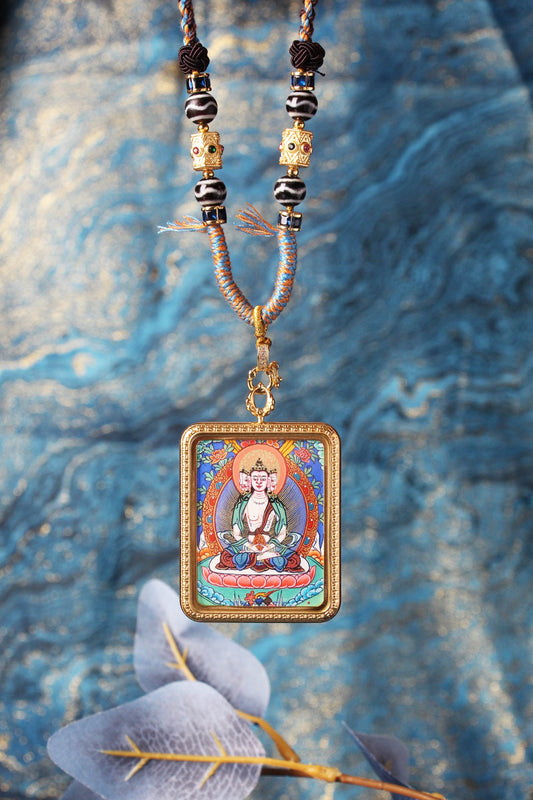 Close-up of a hanging Vairocana Buddha mini thangka pendant in a gold-tone titanium steel case, with braided cord and beadwork against a blue textured backdrop.