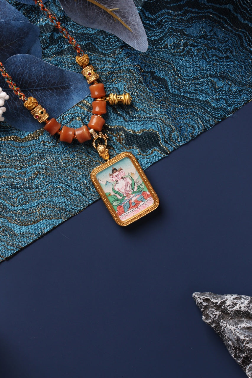 Vairocana Buddha mini thangka pendant necklace showing a hand-painted multi-faced deity seated on a lotus base inside a rectangular metal frame, with braided cord and orange bead accents