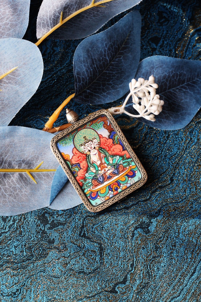 Angled view of a Vairocana Buddha thangka pendant from Rebgong, mineral pigment painting in a silver case, placed on blue patterned fabric with decorative leaves.
