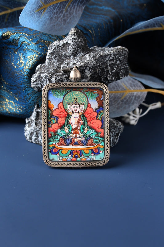 Close-up of a Vairocana Buddha thangka pendant hand-painted by a lama artist in Rebgong, using mineral pigments, set in a silver case, displayed against rock and blue fabric backdrop.
