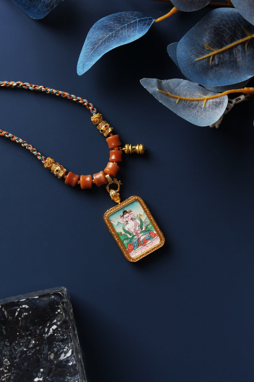 Vairocana Buddha thangka pendant necklace laid flat, featuring a braided cord, orange beads, metal bead components, and a small prayer wheel charm beside the rectangular pendant