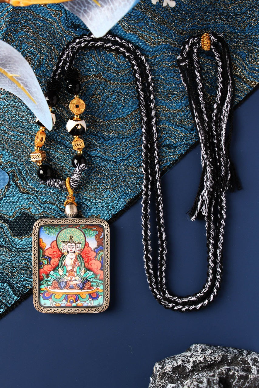 Hand-painted Vairocana Buddha thangka pendant necklace from Rebgong (Regong), mineral pigments, silver case, black-and-white braided cord with beads, flat lay on blue textile background.