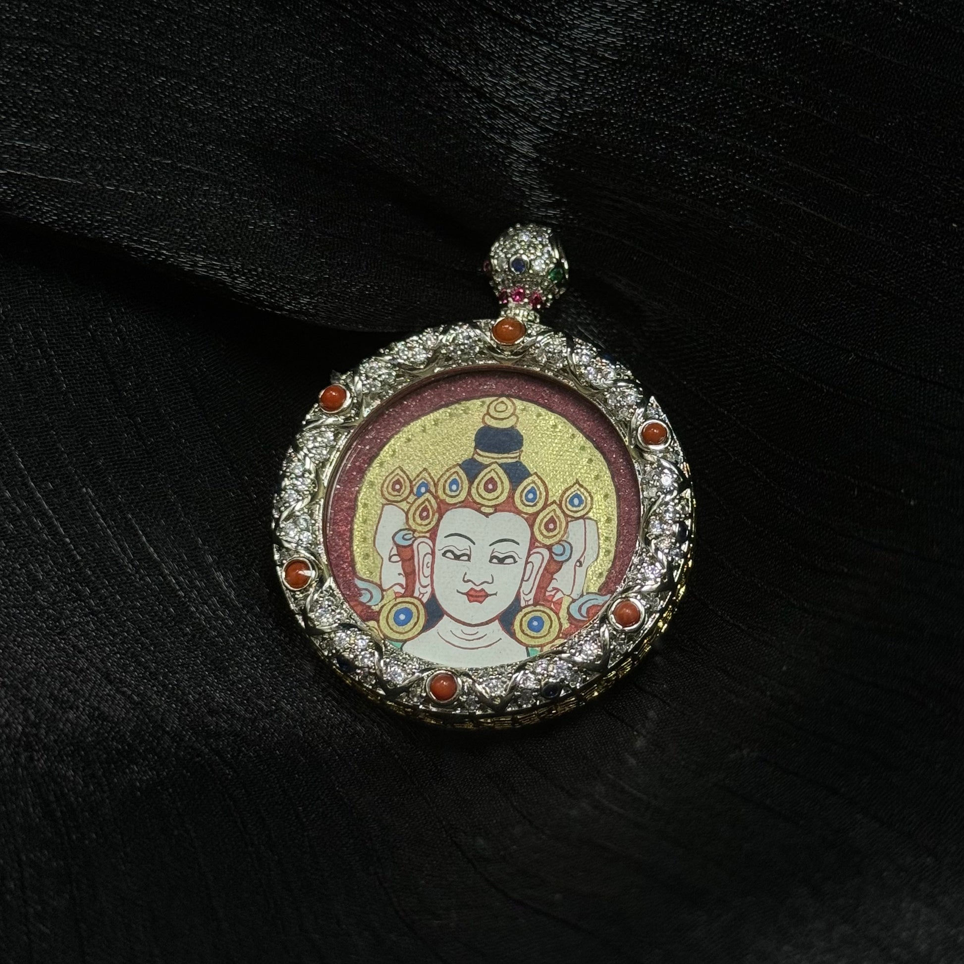 Front view of Vairocana Buddha miniature thangka pendant resting on black silk fabric showing traditional Rebgong hand-painted Buddha head and ornate copper pendant setting