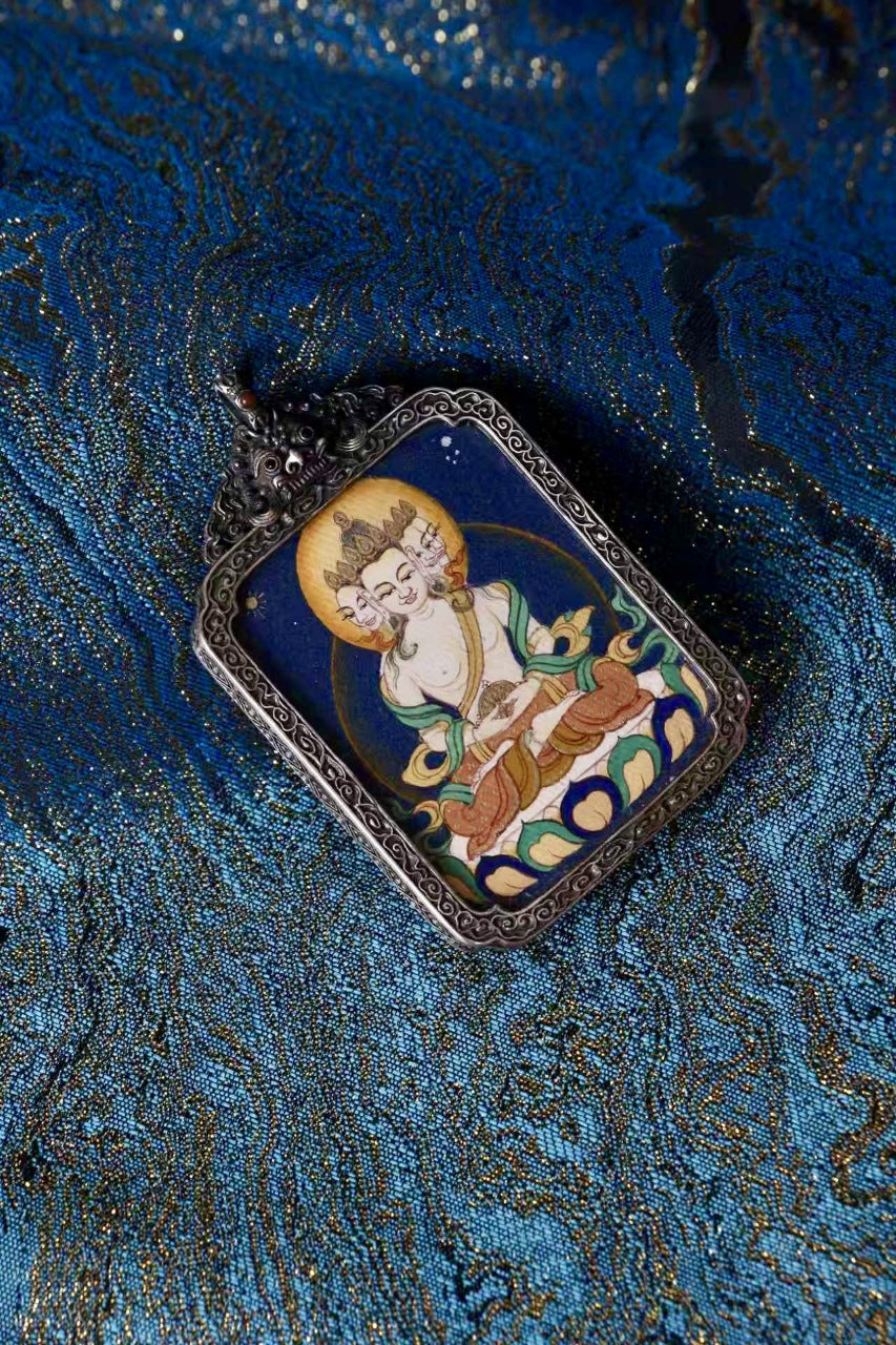 Close-up of a Vairocana (Mahavairocana) mini thangka pendant in an antique silver case on blue brocade fabric, highlighting mineral pigment painting details.