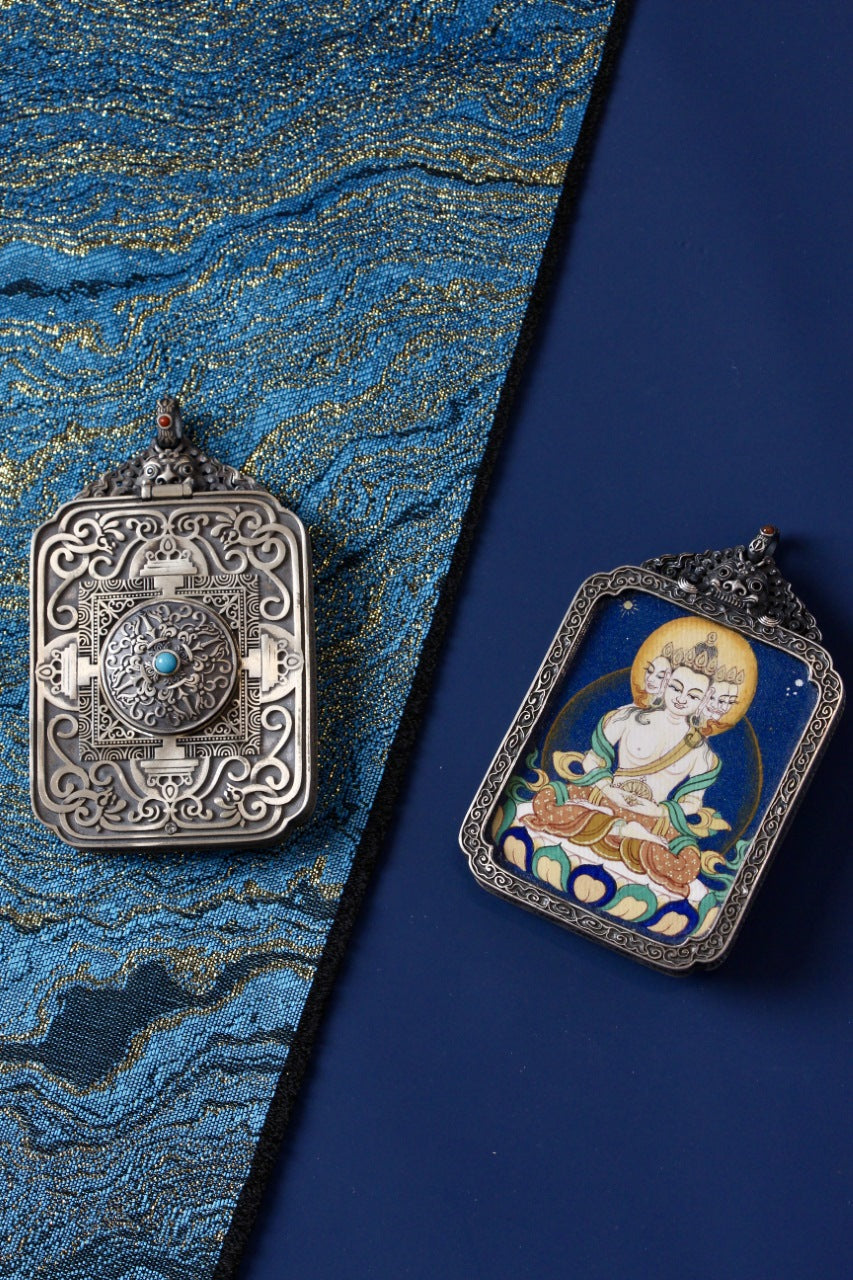 Vairocana mini thangka pendant shown with its ornate antique silver backplate featuring a turquoise accent, on a blue brocade background.