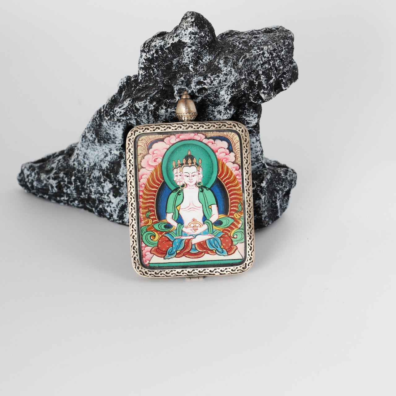 Front view of Vairocana mini Thangka pendant, hand-painted with mineral pigments, showing a multi-faced Buddha depiction, turquoise-green halo, pink cloud background, and an engraved pure silver frame.