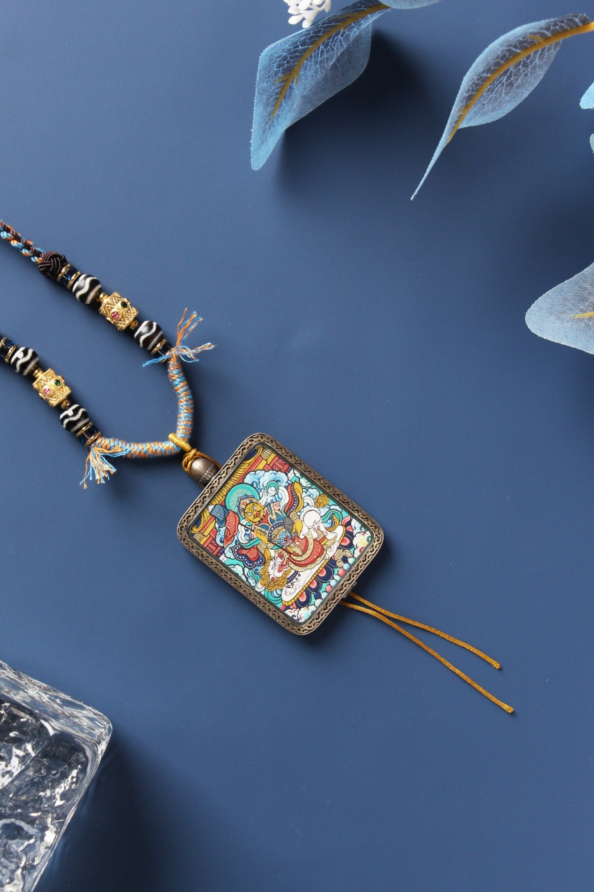 Angled flat-lay of Vaishravana (Bishamonten) mini thangka pendant, hand-painted with mineral pigments in Rebgong style, set in a Dunhuang Nine-Color Deer sterling silver protective case with braided cord and tassel ends