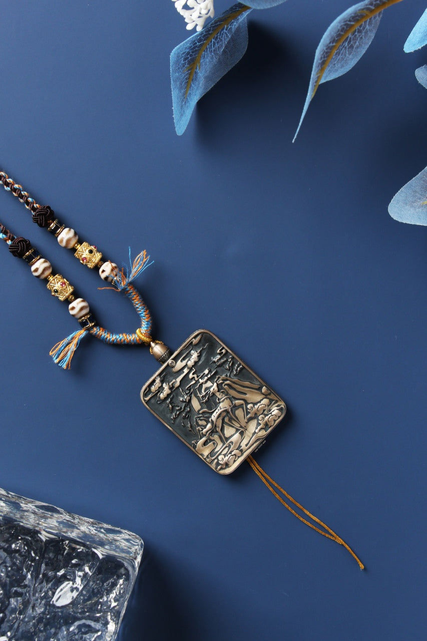 Back view of the Vaishravana mini thangka pendant showing an embossed sterling silver case with sculpted relief details, attached braided cord, and double tassel ends in a minimalist studio flat-lay