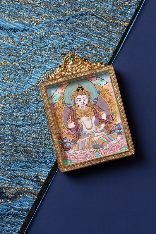 Angled flat lay of Vajrasattva mini thangka pendant, hand-painted mineral pigment artwork in a jeweled double-sided titanium steel case, Nepal high-end series