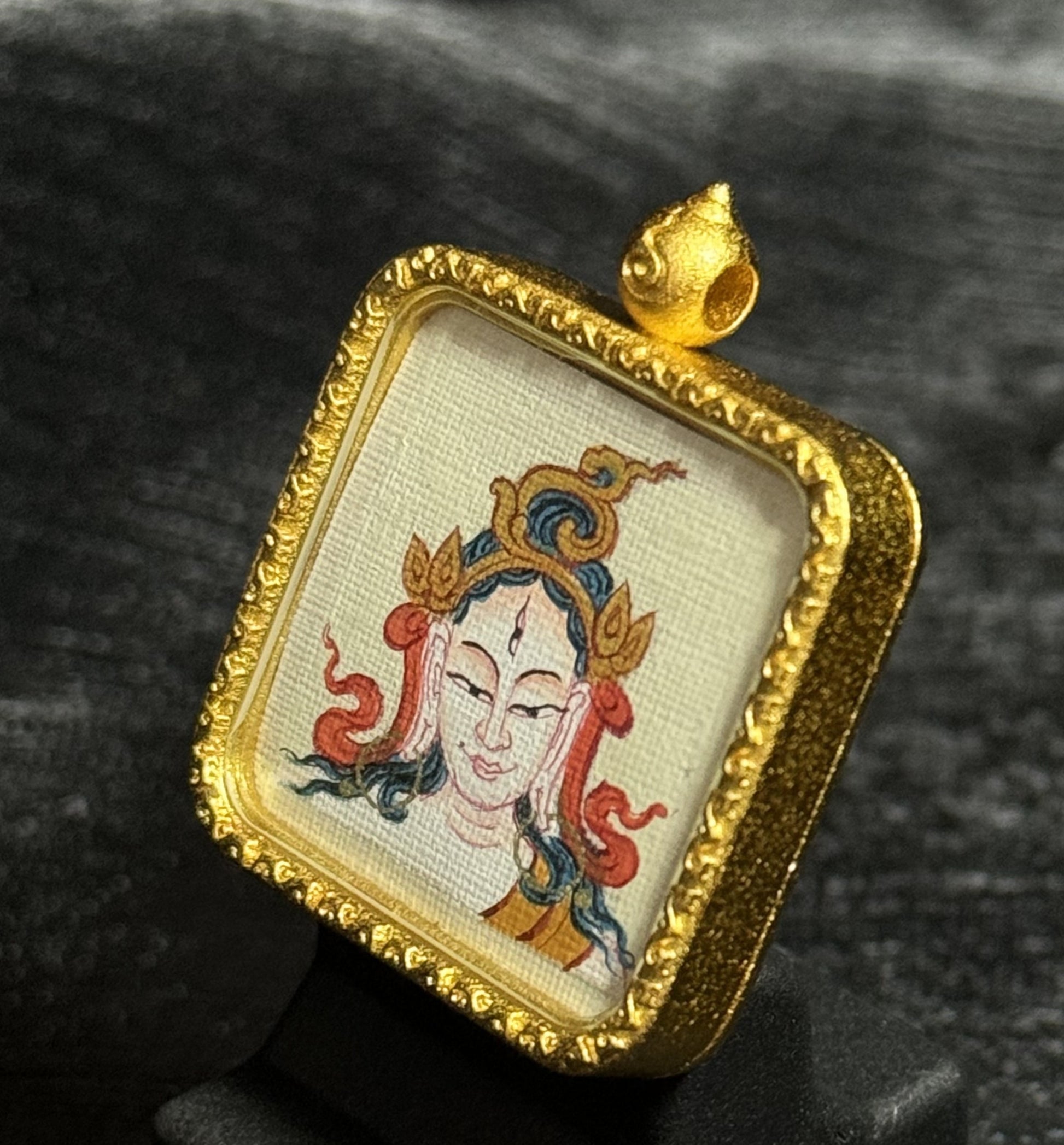 Angled view of a White Tara mini thangka pendant showing the hand-painted deity image on textured canvas, framed by an ornate gold-toned pendant setting.