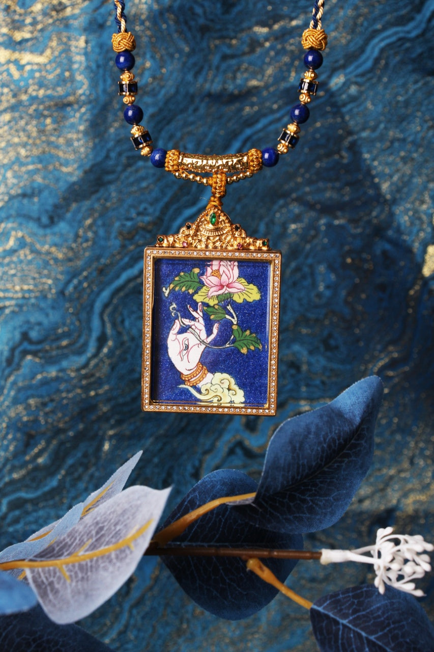 Front view of White Tara hand thangka pendant necklace, lotus and offering hand painted on rich blue, gold-tone double-layer jeweled frame, crown top with green accent stone and blue bead chain.