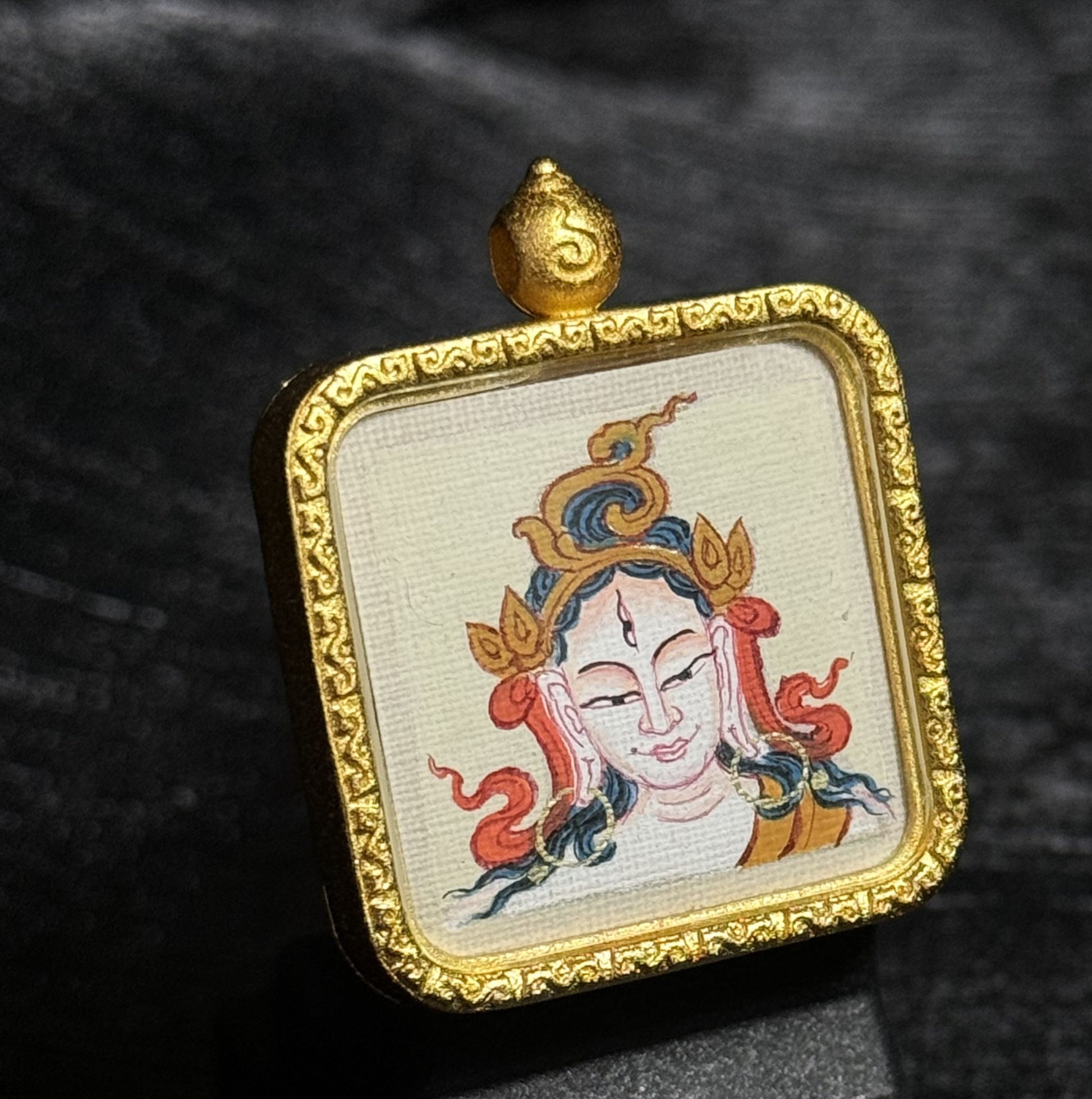White Tara miniature thangka pendant displayed on dark fabric background, showing hand-painted Tibetan deity portrait within a decorative gold-toned pendant frame.