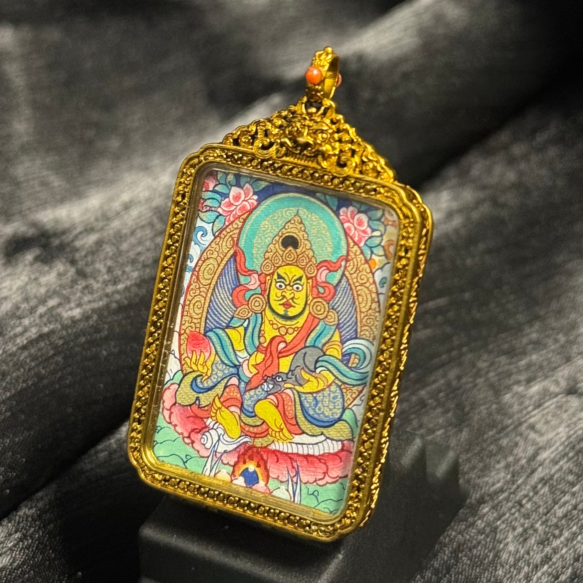 Hand-painted Yellow Jambhala mini thangka pendant placed on a display stand, showing the wealth deity holding a jewel and mongoose, surrounded by decorative floral and halo patterns within a gold copper frame.