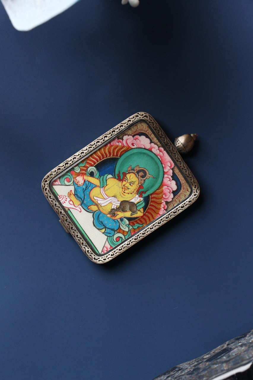 Angled product view of the Yellow Jambhala mini thangka pendant from the Tibetan Red Cloud Zangka Series, fully hand-painted in Tibet with natural mineral pigments and presented in a Dunhuang Nine-Colored Deer sterling silver case for portable devotion and collecting.