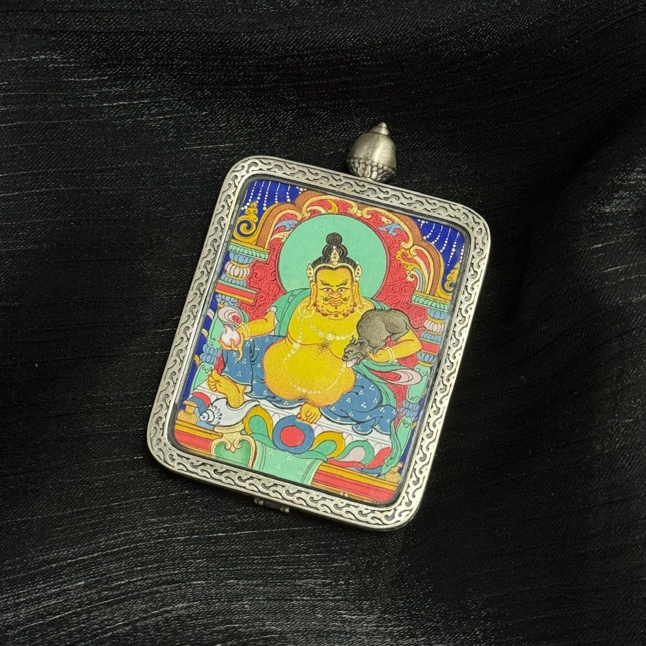 Angled view of Yellow Jambhala mini thangka pendant, highlighting the silver gawu frame, hand-painted artwork surface, and balanced Tibetan composition.