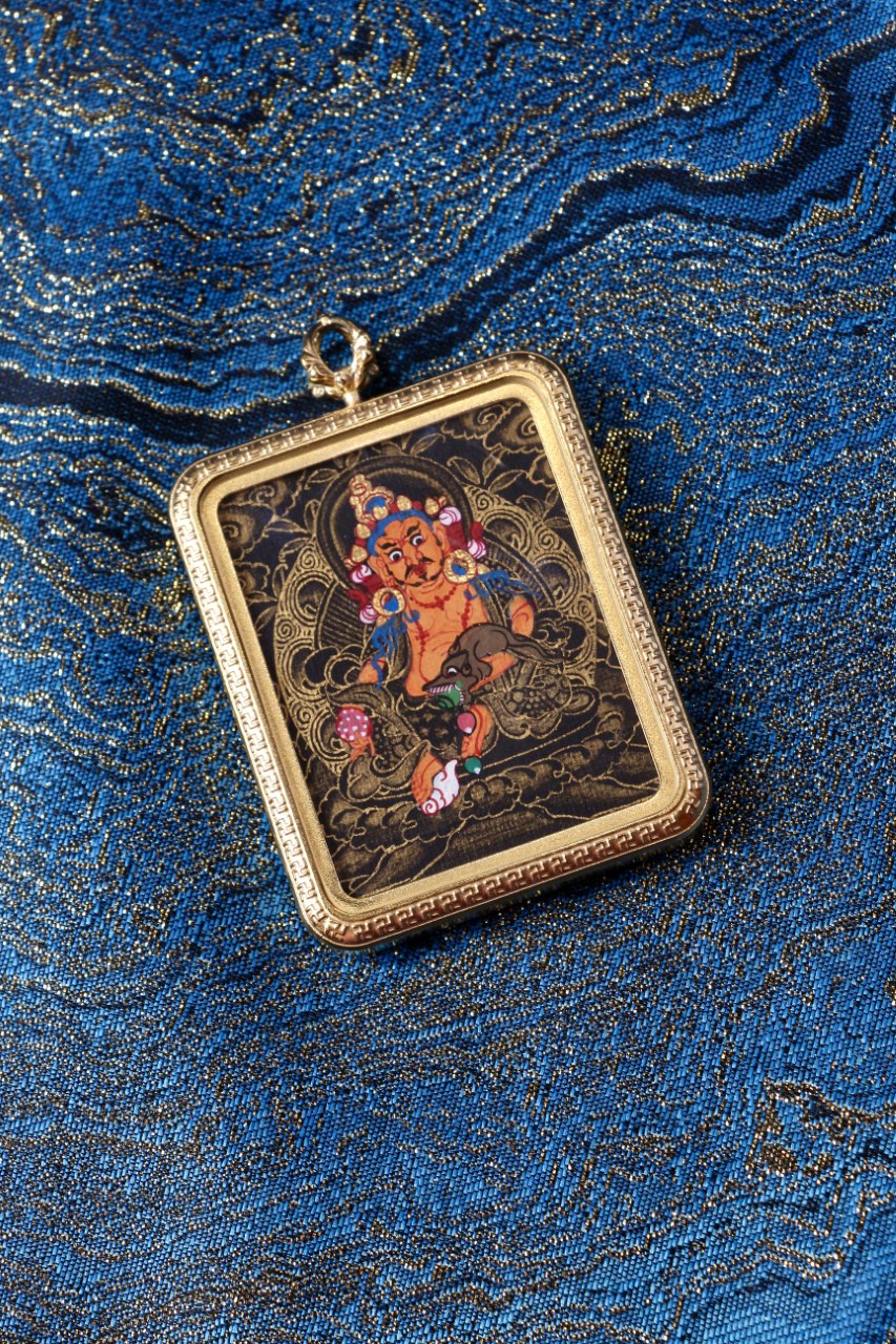 Close-up of Yellow Jambhala black-gold miniature thangka pendant in a gold-tone titanium steel case, photographed on traditional blue brocade fabric
