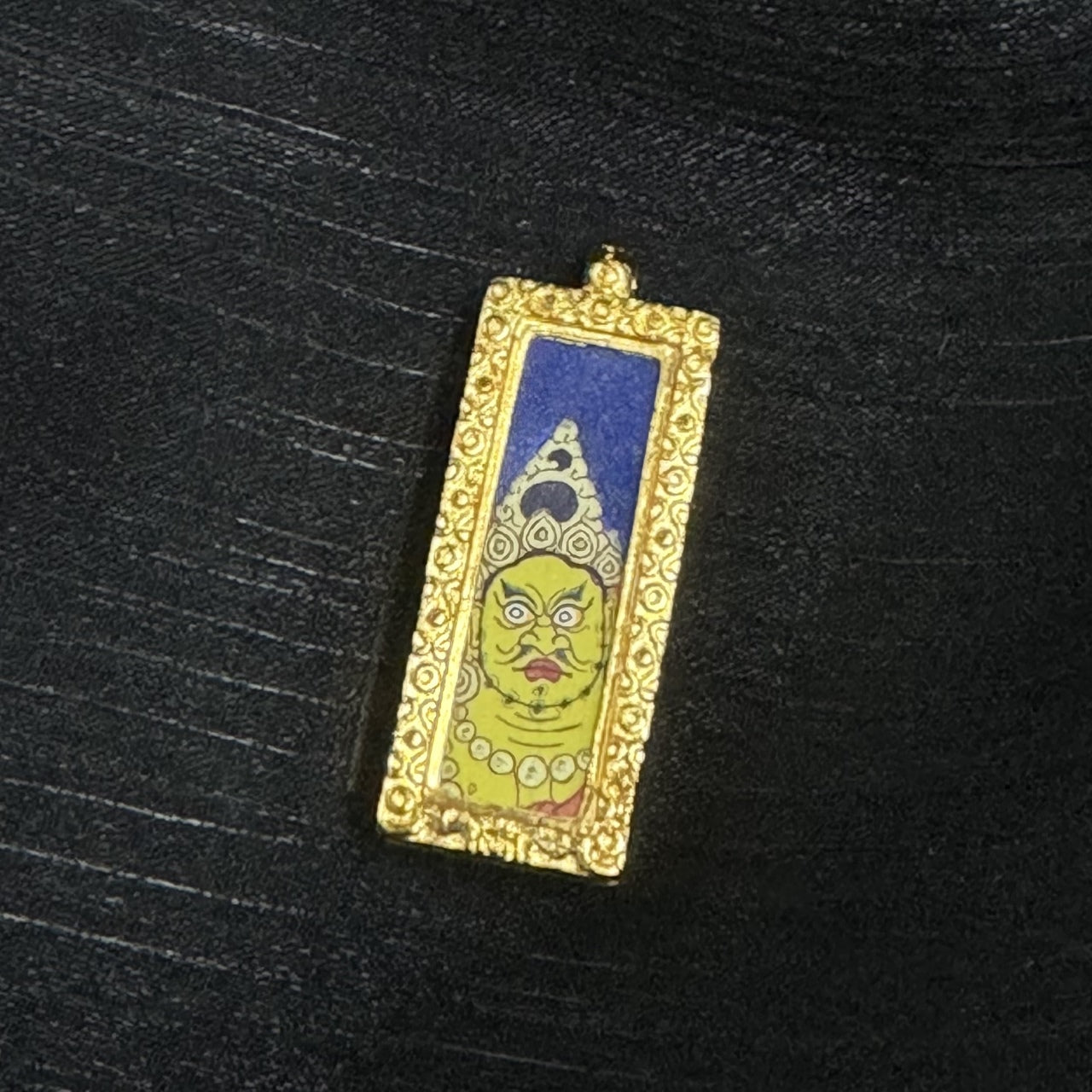 Close-up of the Yellow Jambhala mini thangka pendant highlighting fine linework in the eyes and brows, layered mineral-pigment color blocks, and the textured ornamental border on the pendant frame.