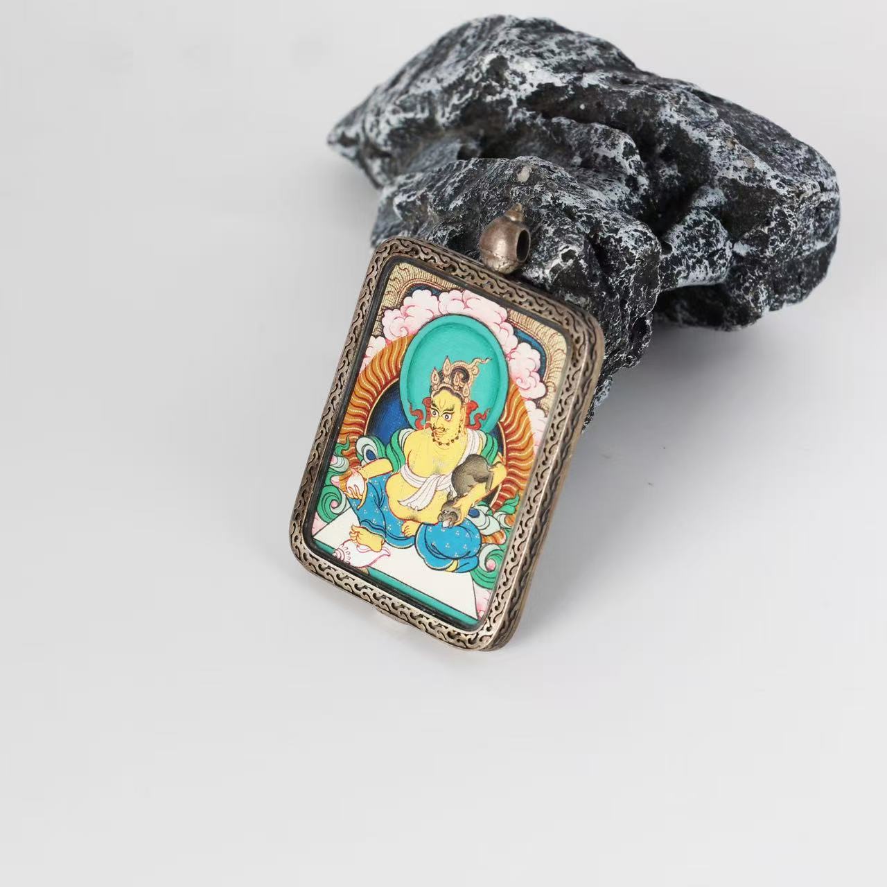 Close-up of Yellow Jambhala mini thangka pendant showing fine line work, cloud motifs, mineral pigment texture, and the engraved pattern around the sterling silver case