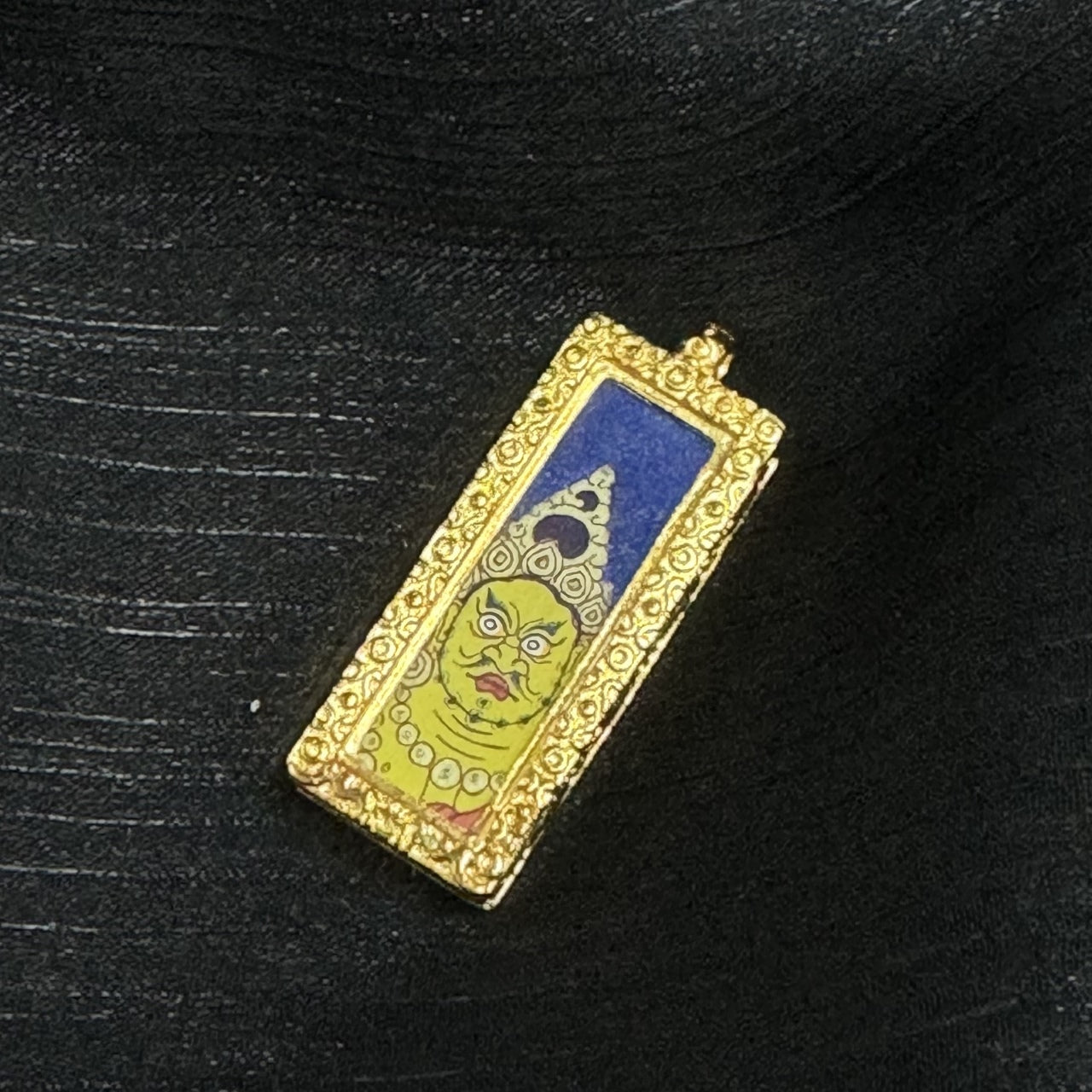 Front view of the Yellow Jambhala mini thangka pendant showing the tall rectangular format, deep blue upper background, crown motif details, and the hand-painted yellow face framed by a raised ornamental border.