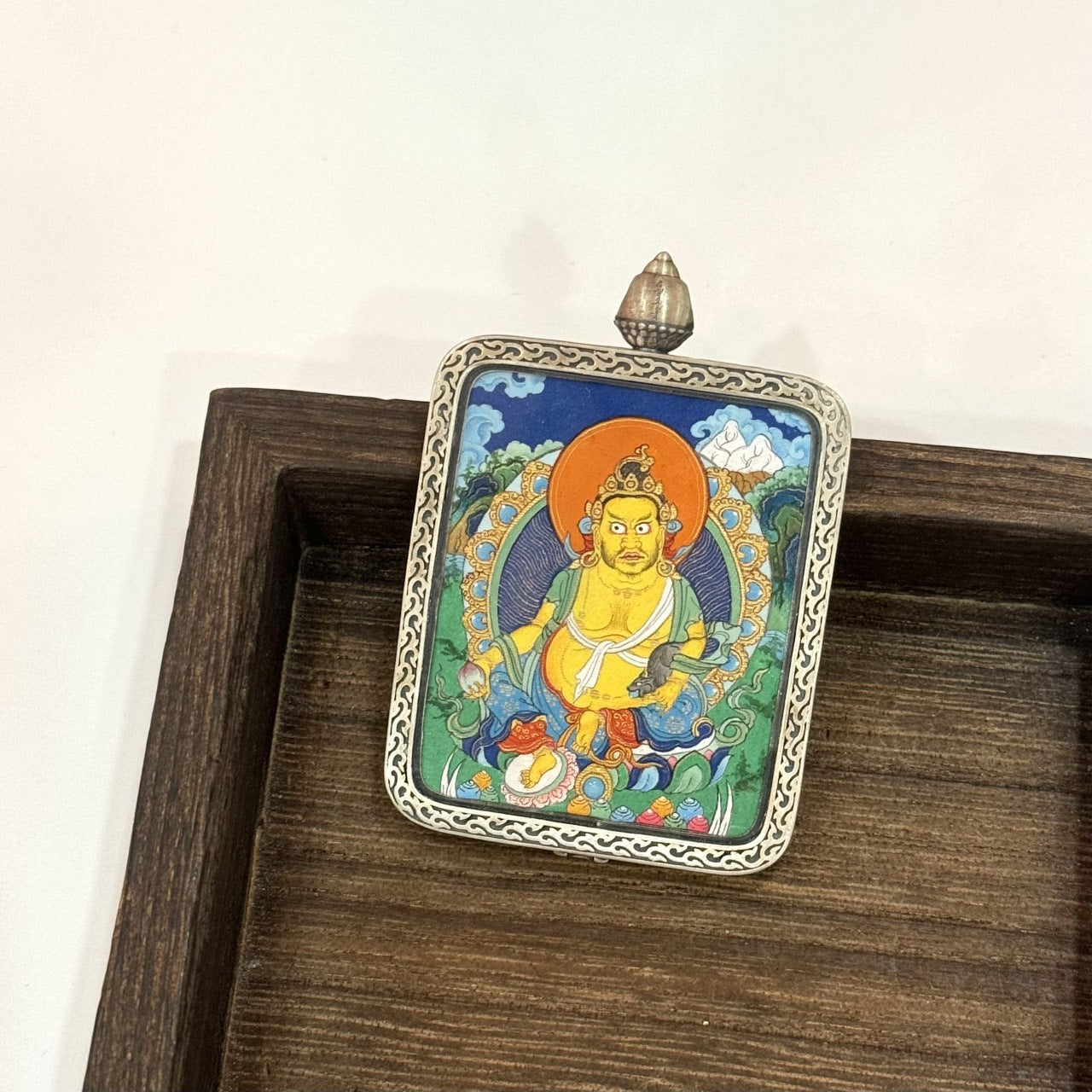 Front view of Yellow Jambhala mini thangka pendant, hand-painted with mineral pigments, set in a Dunhuang Nine-Color Deer silver case with engraved border and top bail.