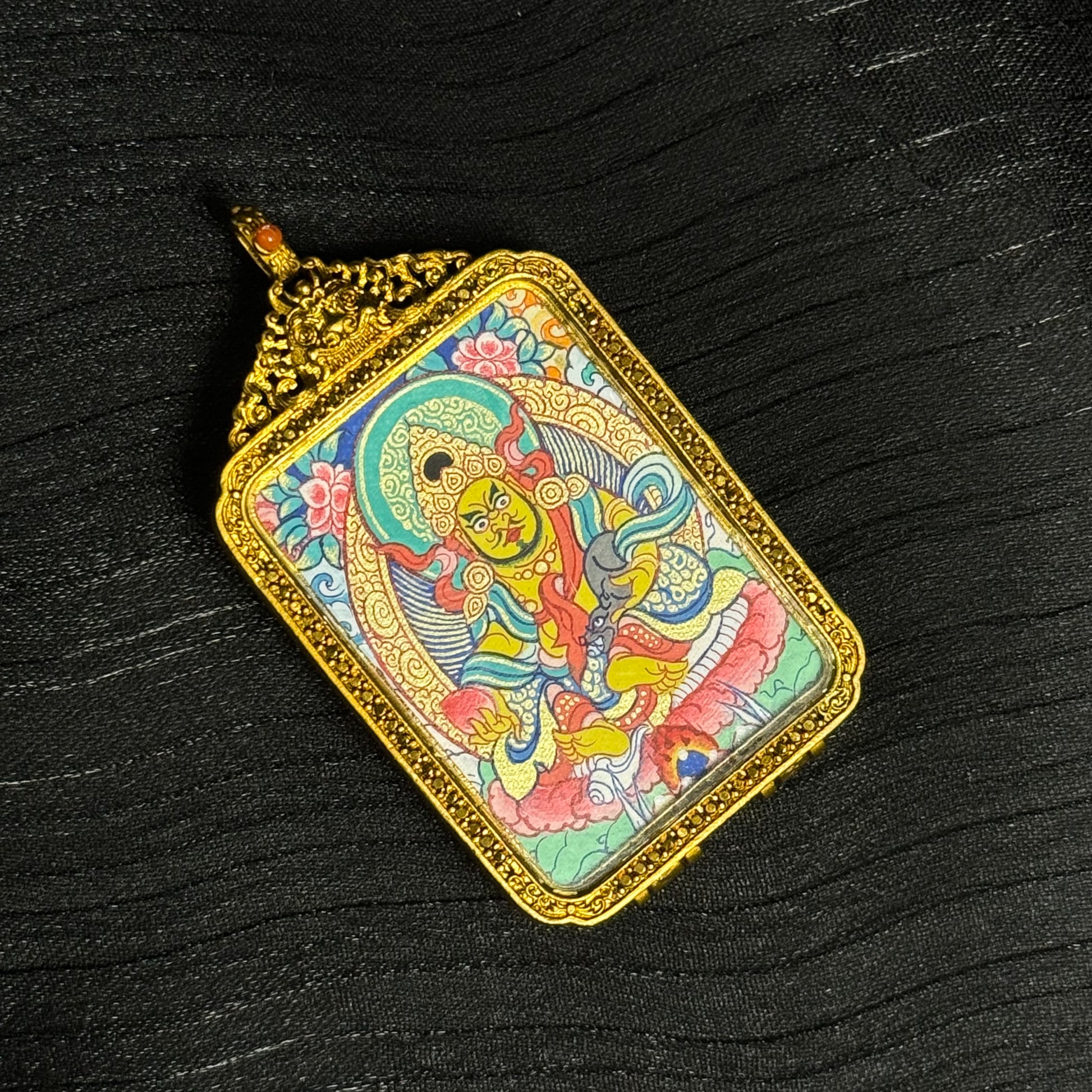 Portable Yellow Jambhala mini thangka pendant photographed on dark fabric background, highlighting the detailed Tibetan iconography, mineral pigment painting, and ornate gold copper protective case.