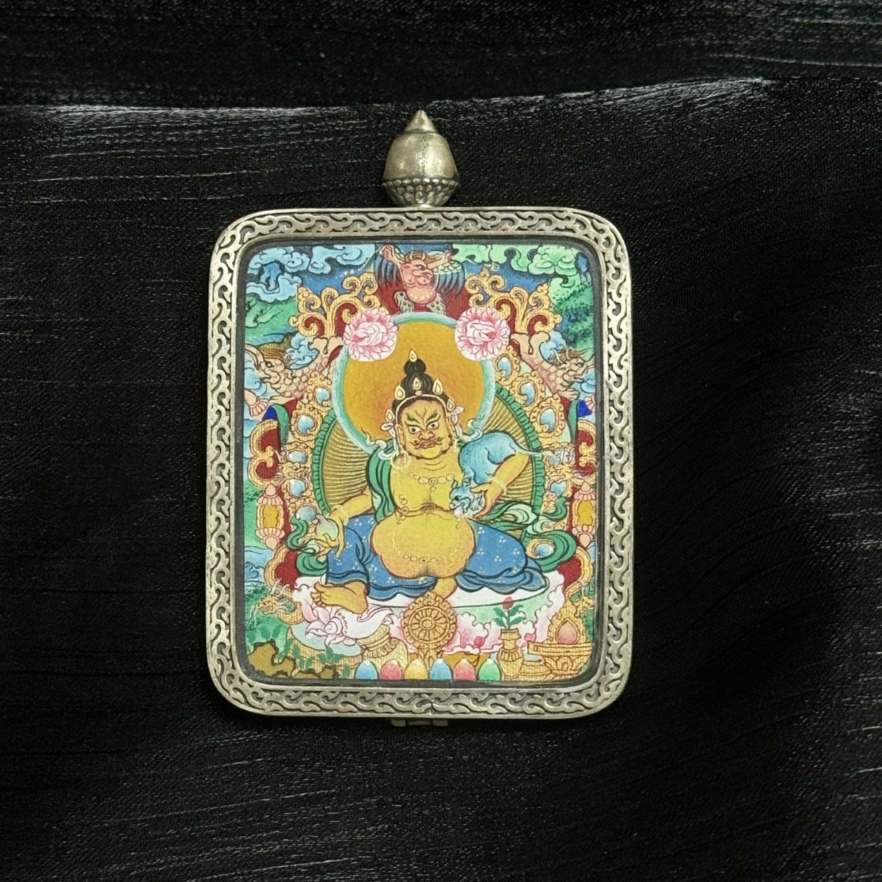 Full frontal composition of Yellow Jambhala mini thangka pendant in silver Dunhuang Nine-Color Deer case, displaying detailed deity posture, wealth symbols, decorative floral motifs, and textured mineral pigments.