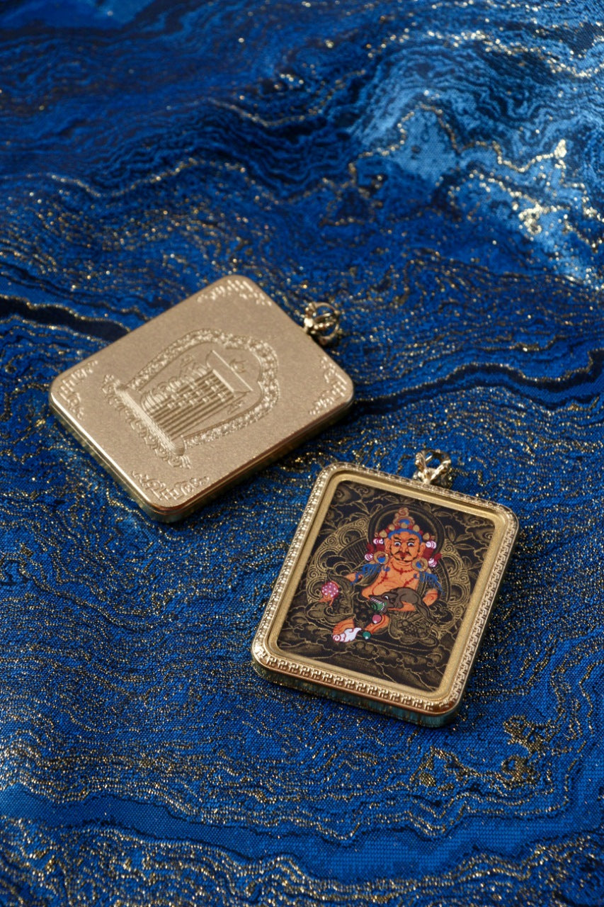 Yellow Jambhala mini thangka pendant shown with the protective case, including front black-gold painting and back-side metal plate detail on blue brocade fabric