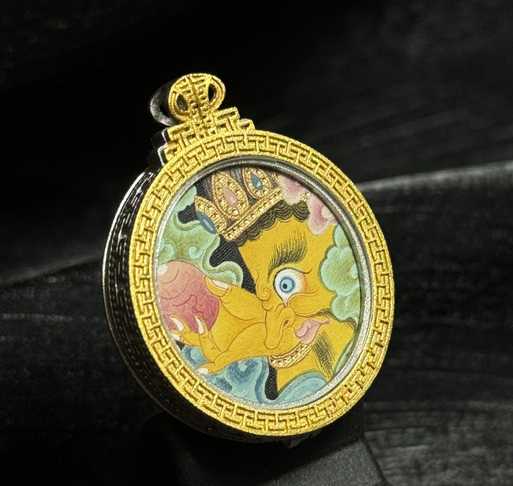 Side-angle view of the Yellow Jambhala mini thangka pendant showing the circular gold-toned frame, hand-painted deity artwork, and the thickness of the protective silver pendant case.