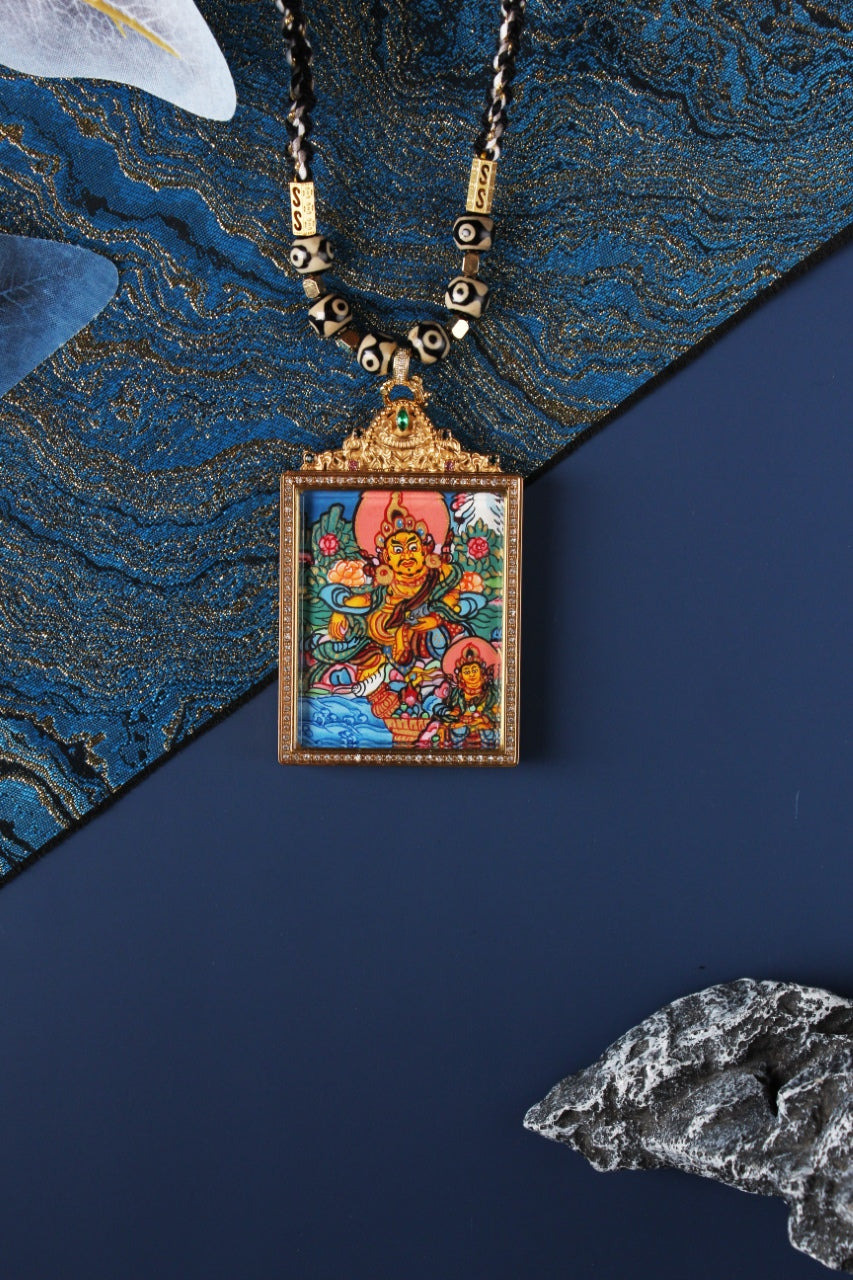 Front view of a hand-painted dual-deity thangka pendant featuring Yellow Jambhala with a smaller accompanying deity, set in a square jeweled frame with crystal border and ornate top crown with green stone.