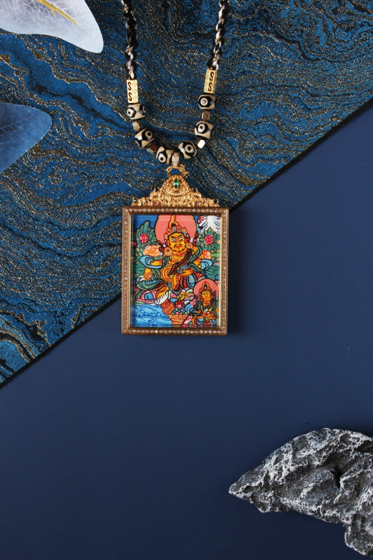 Front view of a hand-painted dual-deity thangka pendant featuring Yellow Jambhala with a smaller accompanying deity, set in a square jeweled frame with crystal border and ornate top crown with green stone.