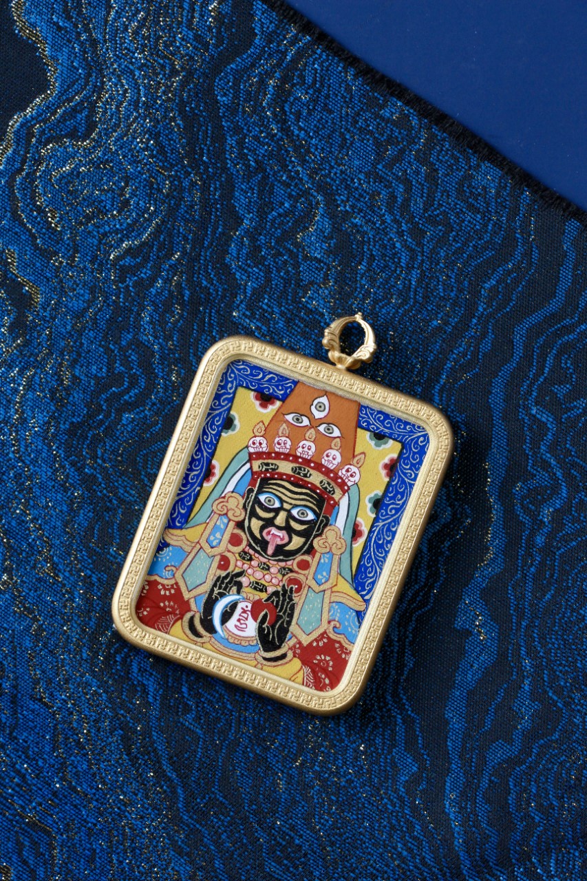 Front view of Zaki Lhamo mini thangka pendant on blue background, fully hand-painted with mineral pigments in a gold-tone copper frame.