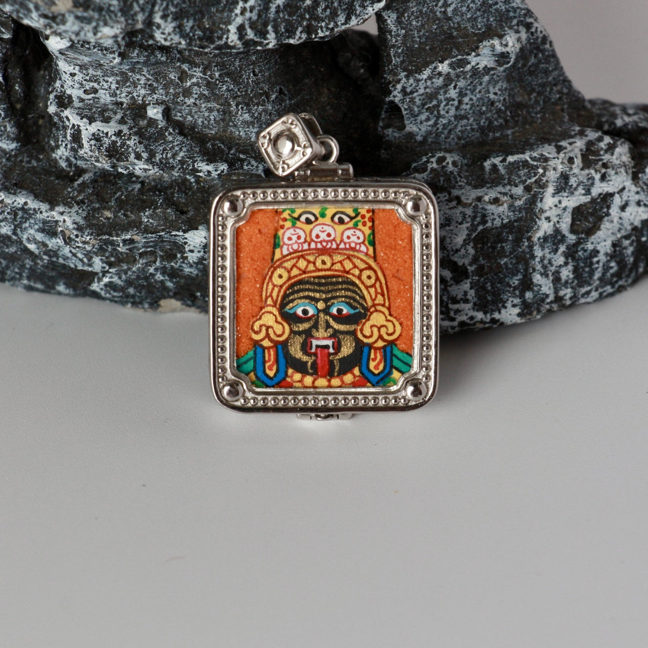 Front close-up of Zaki Lhamo mini thangka pendant with hand-painted mineral pigments, orange background, and detailed facial features in a square silver-plated frame.