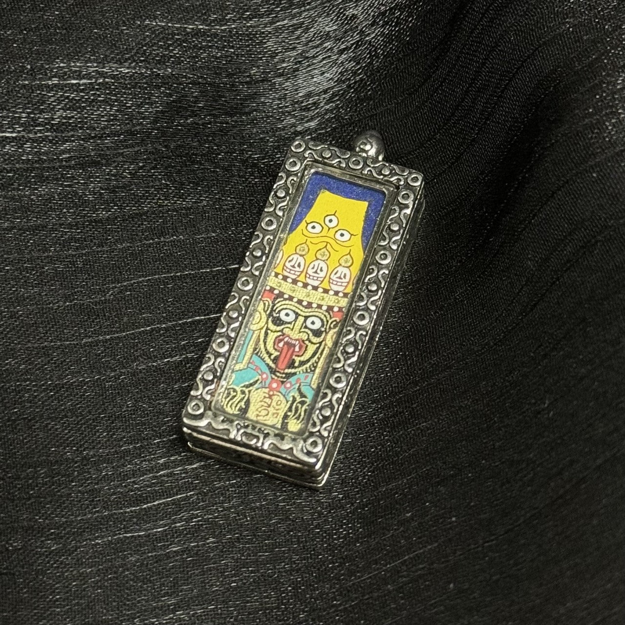 Angled front view of Zaki Lhamo amulet pendant featuring a fierce deity face painting, handmade with mineral pigments in Rebgong style, set in a metal pendant case.
