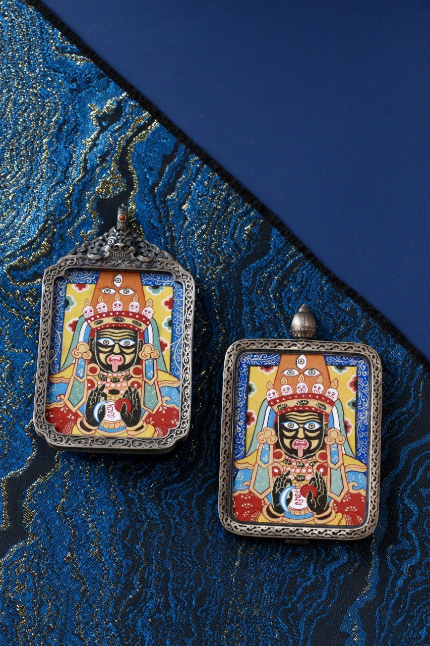 Two Zaki Lhamo hand-painted mini thangka pendants in 925 silver cases, shown side by side on blue brocade backgroundpigments.