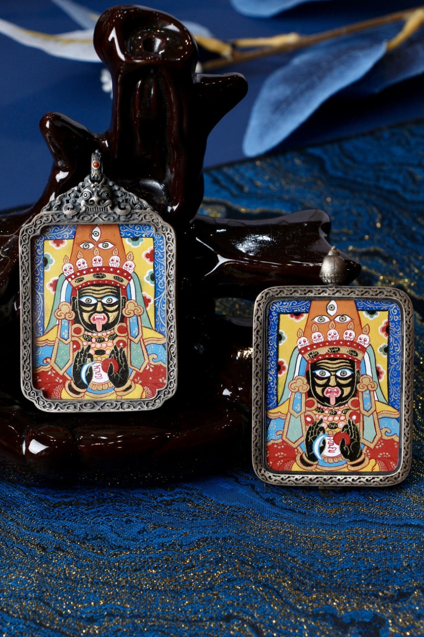 Two Zaki Lhamo hand-painted mini thangka pendants in different 925 silver cases, staged on a dark sculpture with blue textile backdrop