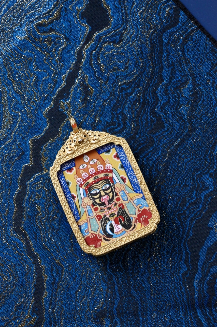 Ornate-style Zaki Lhamo gawu pendant shown at an angle, featuring a sculpted gold-tone copper frame and hand-painted mineral pigment thangka.
