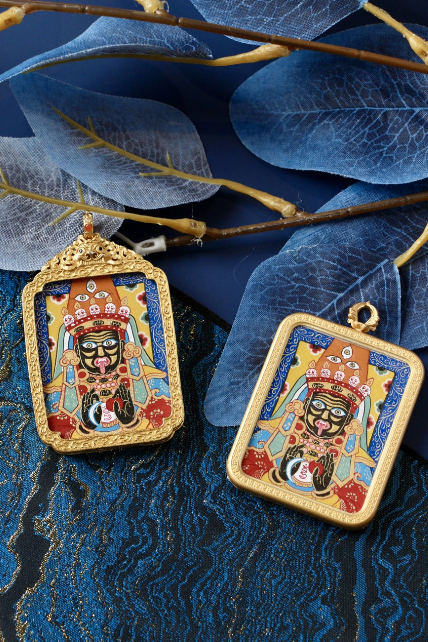 Two Zaki Lhamo mini thangka pendants photographed with decorative leaves, highlighting the ornate gawu frame and the classic square frame in gold-tone copper.