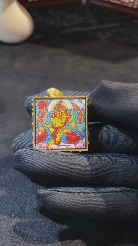 Close-up video of a double-sided hand-painted Yellow Jambhala mini Thangka pendant, featuring the deity holding the treasure-spitting mongoose in an 18K gold Gau box with sapphire crystal