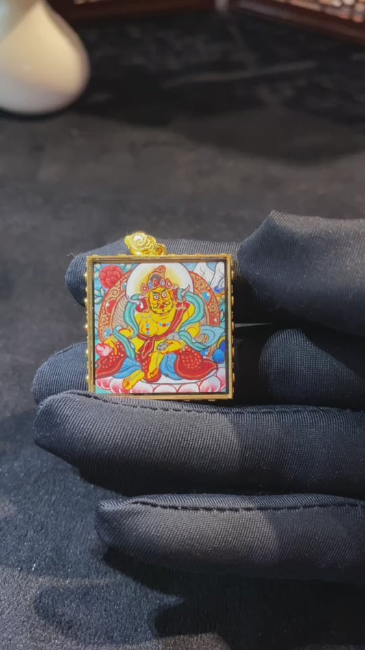 Close-up video of a double-sided hand-painted Yellow Jambhala mini Thangka pendant, featuring the deity holding the treasure-spitting mongoose in an 18K gold Gau box with sapphire crystal